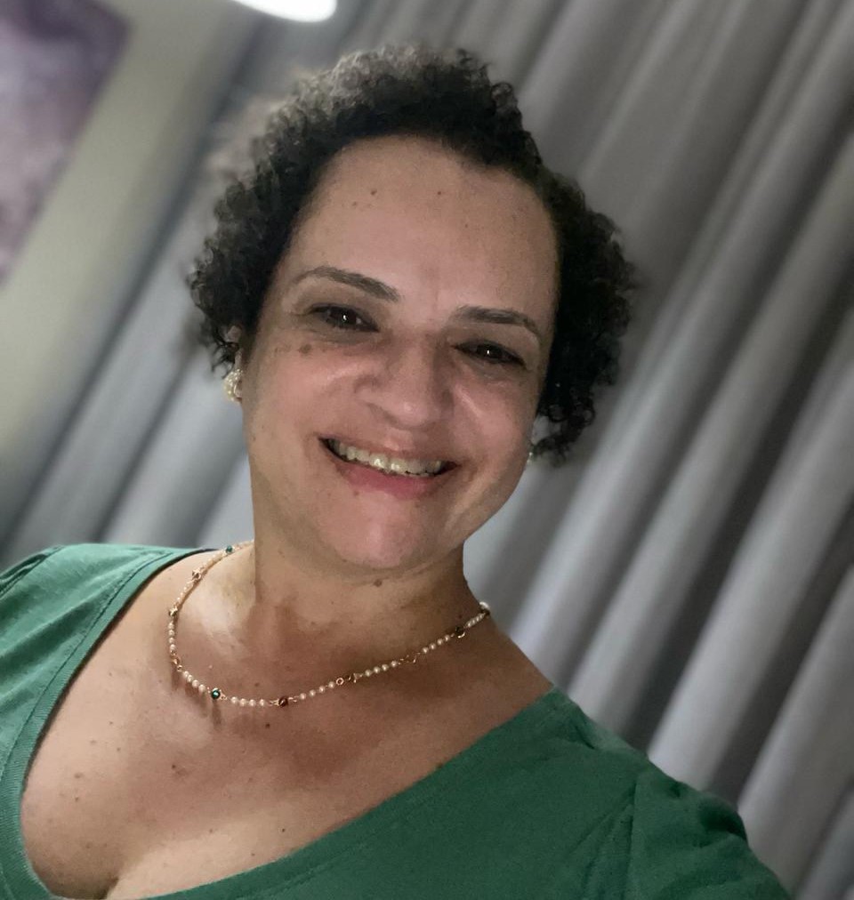 A imagem é a foto de Regiane Fakouri. Ela é uma mulher de pele branca e cabelos pretos, curtos e enrolados . Ela está sorrindo, usa um colar e uma blusa verde.